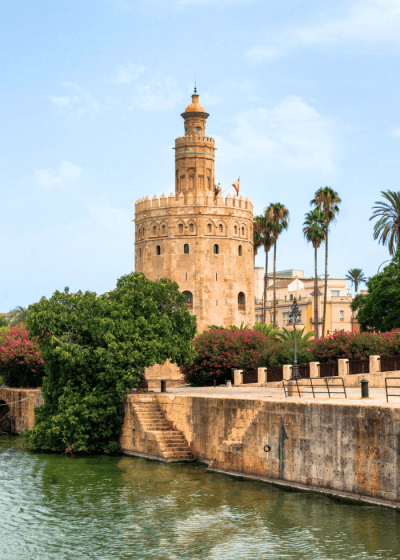 Golden Tower by river surrounded by greenery and palm trees.