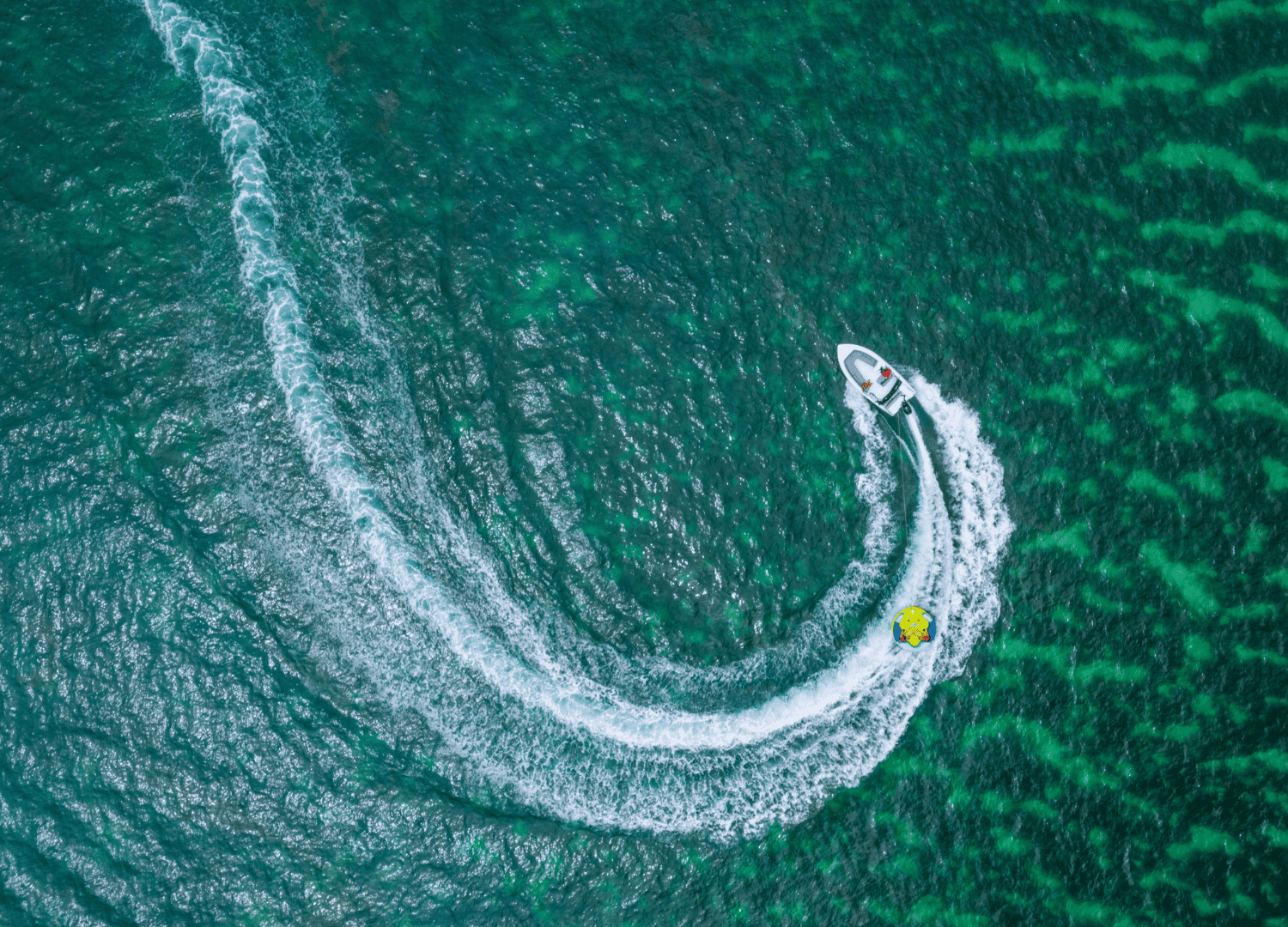 Speedboat towing inflatable on turquoise water, aerial view.